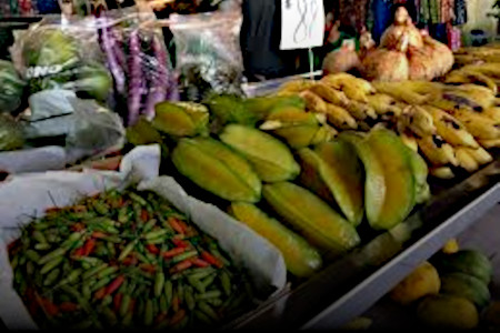 Photo of a produce stall at a street market, offering red and green chiles, starfruit, bananas, coconuts, and other fruits and vegetables.